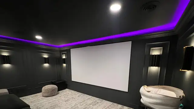 Basement home theater with black walls, a projection screen, wall sconces and purple cove lighting