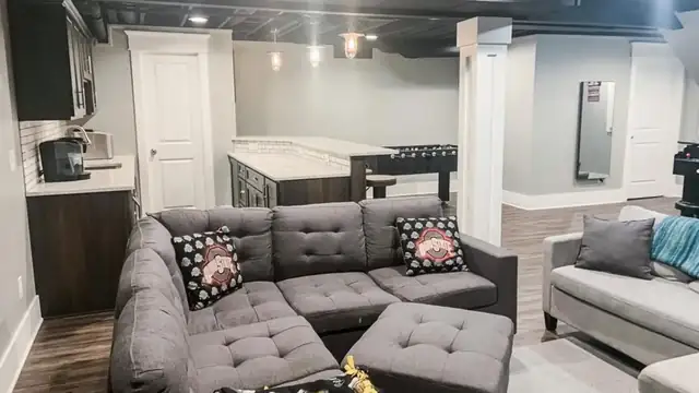 Finished basement lounge with a gray tufted sectional, a kitchenette with dark cabinets and a game area beyond