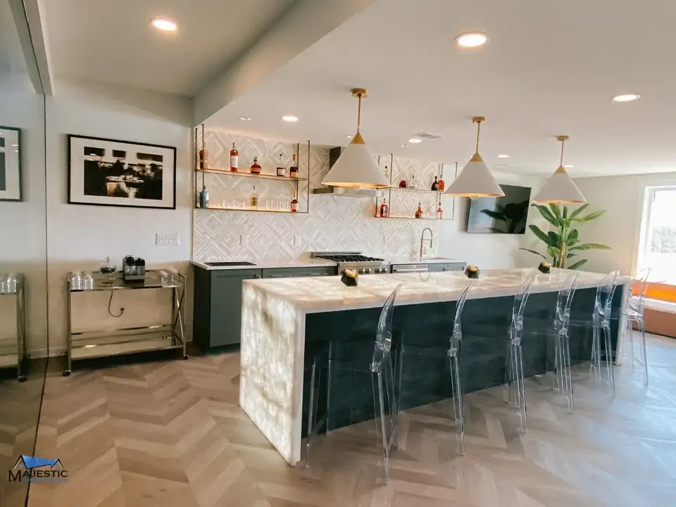 Finished basement bar with a backlit stone-look counter, clear stools and herringbone floor, built by Majestic Home Solutions, a remodeling contractor in Sterling Heights