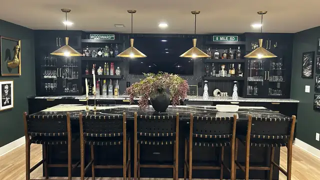 Basement bar with dark green cabinets, glass shelving, brass cone pendants and a row of leather bar stools